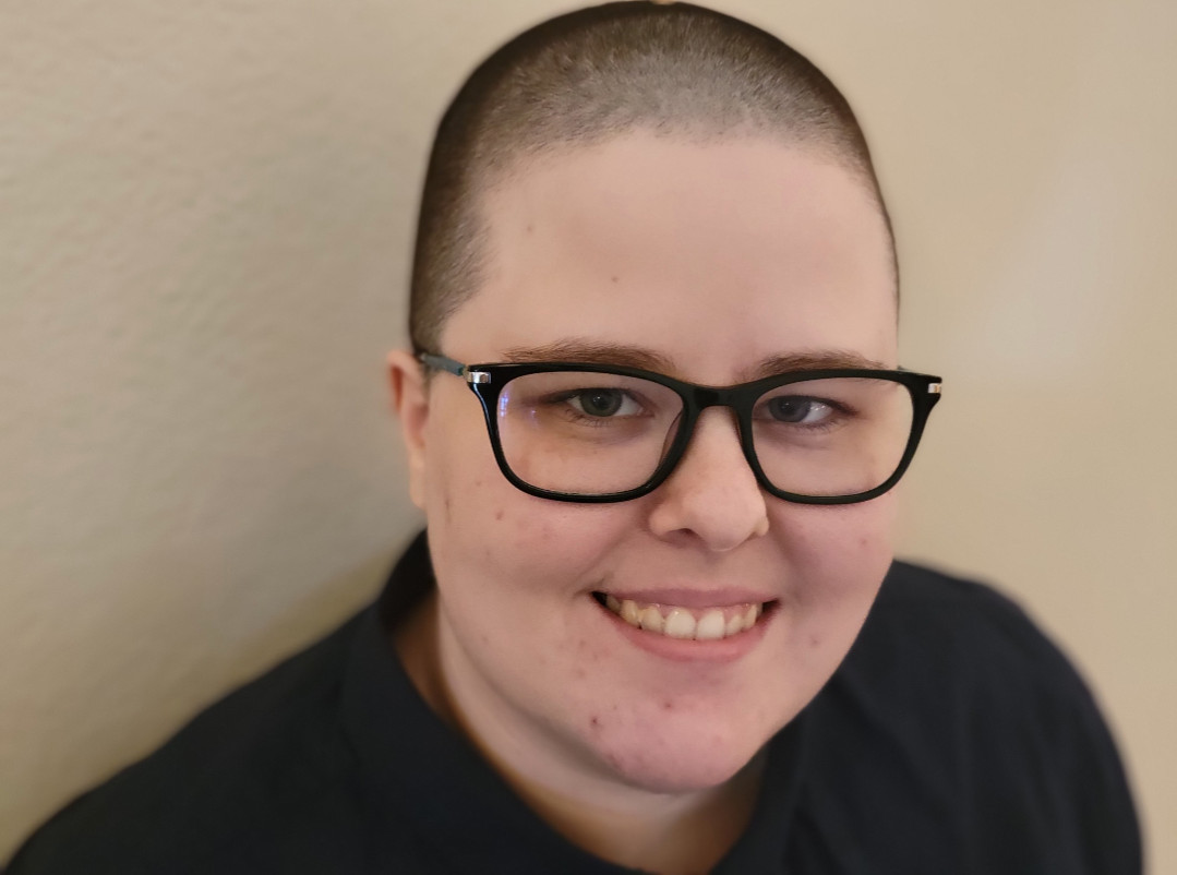 A person with a shaved head and black glasses smiles at the camera. They have a dark blue collared shirt on.